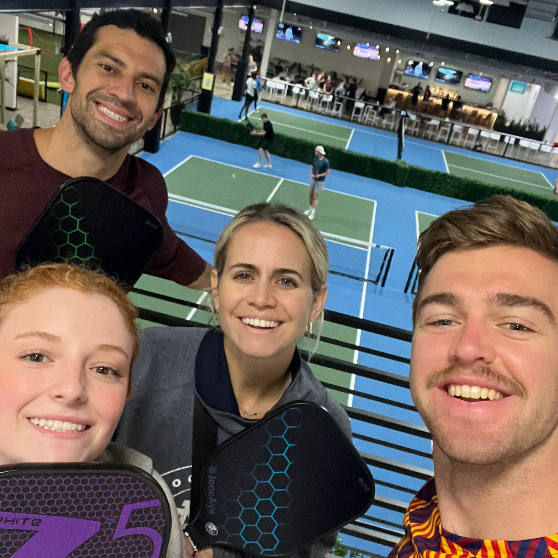 Four young, fun people posing for a selfie with a pickleball court in the background. Four, young cute men posing together on a pickleball court with equipment. Young man playing pickleball on an outdoor court with palm trees in the background, Man playing pickleball on an outdoor court with a fence and trees in the background. Playing in Young woman holding a pickleball paddle on a court with a blurred background. From Silly Pickles pickleball ladder league (the world&