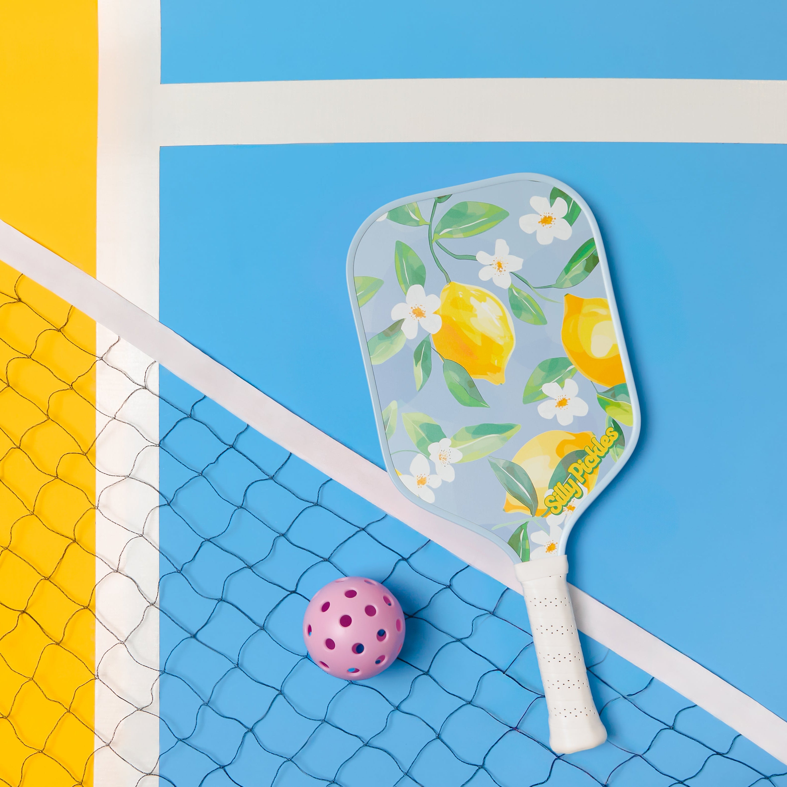 Lemon-patterned Silly Pickles pickleball paddle and pink ball on a blue and yellow court