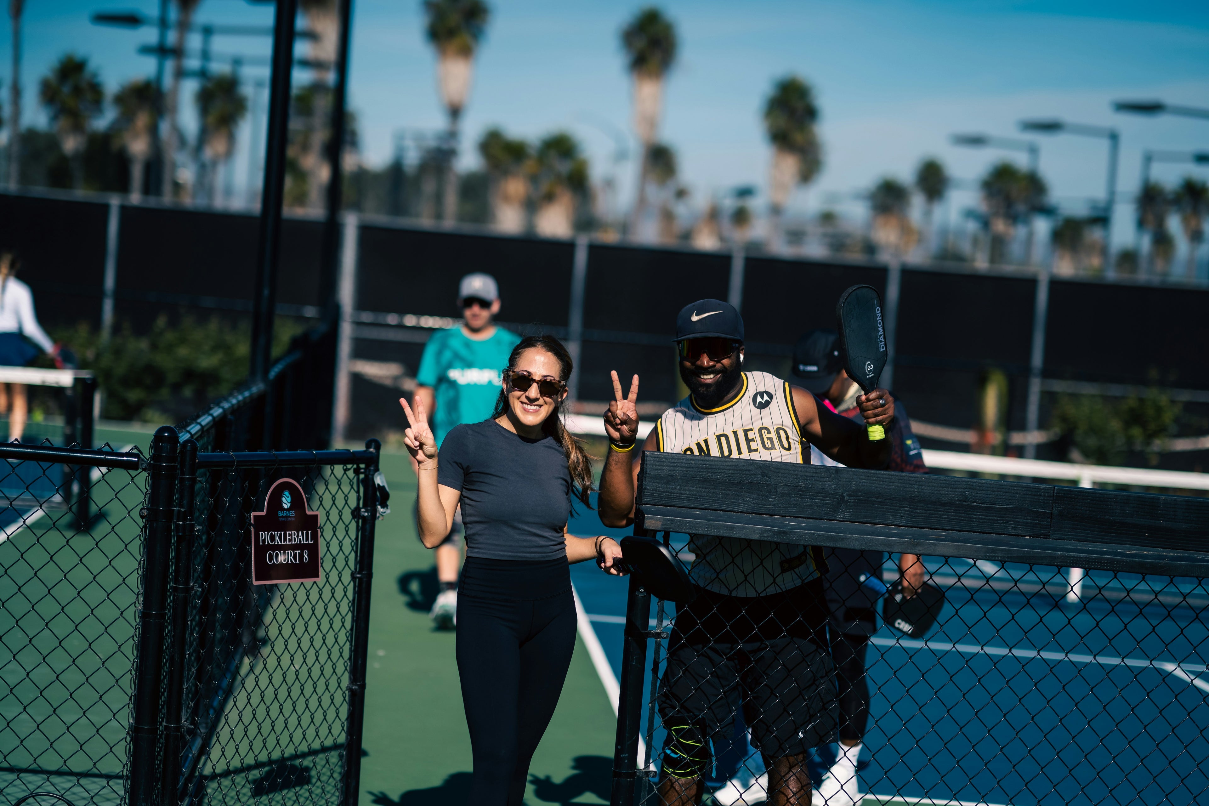 Two young, fun adults on a pickleball court with palm trees in the background. Young man playing pickleball on an outdoor court with palm trees in the background, Man playing pickleball on an outdoor court with a fence and trees in the background. Playing in Young woman holding a pickleball paddle on a court with a blurred background. They are playing in Silly Pickles pickleball ladder league (the world's funnest pickleball league)