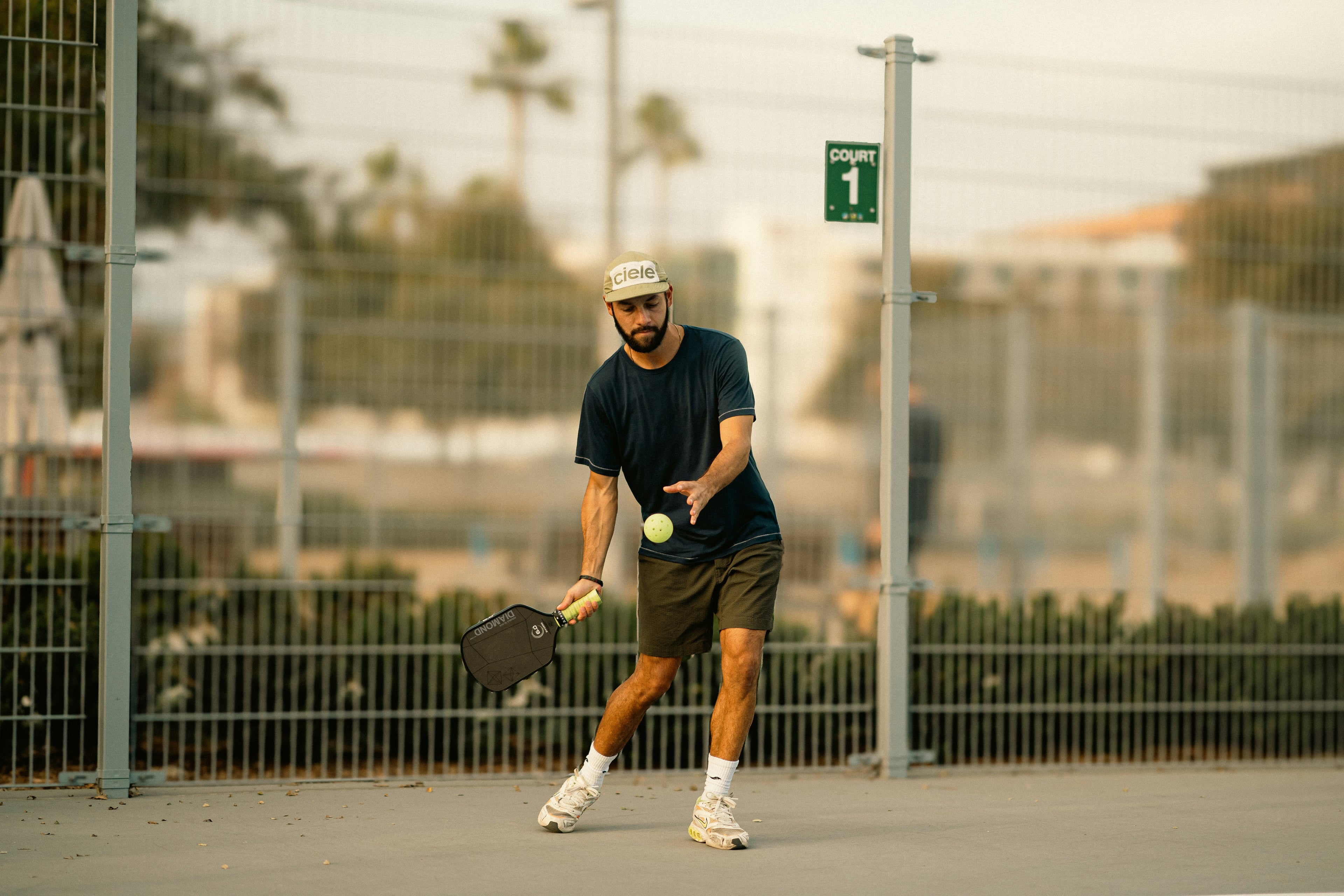 Young man playing pickleball on an outdoor court with palm trees in the background, Man playing pickleball on an outdoor court with a fence and trees in the background. Playing in Young woman holding a pickleball paddle on a court with a blurred background, playing in Silly Pickles pickleball ladder league (the world's funnest pickleball league)