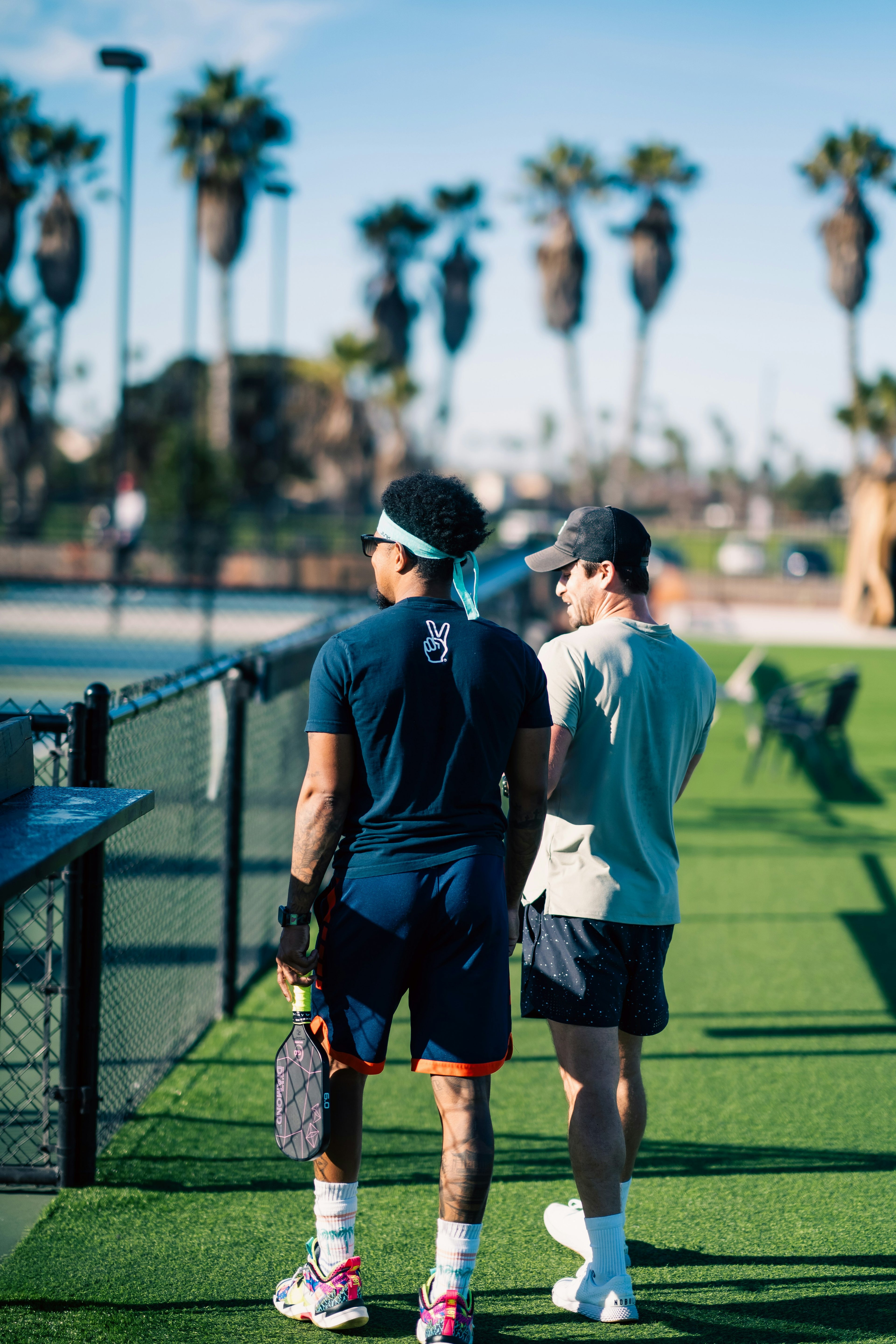 Two pickleball players on a court with palm trees in the background. Young man playing pickleball on an outdoor court with palm trees in the background, Man playing pickleball on an outdoor court with a fence and trees in the background. Playing in Young woman holding a pickleball paddle on a court with a blurred background. Playing in Silly Pickles pickleball ladder league (the world's funnest pickleball league)