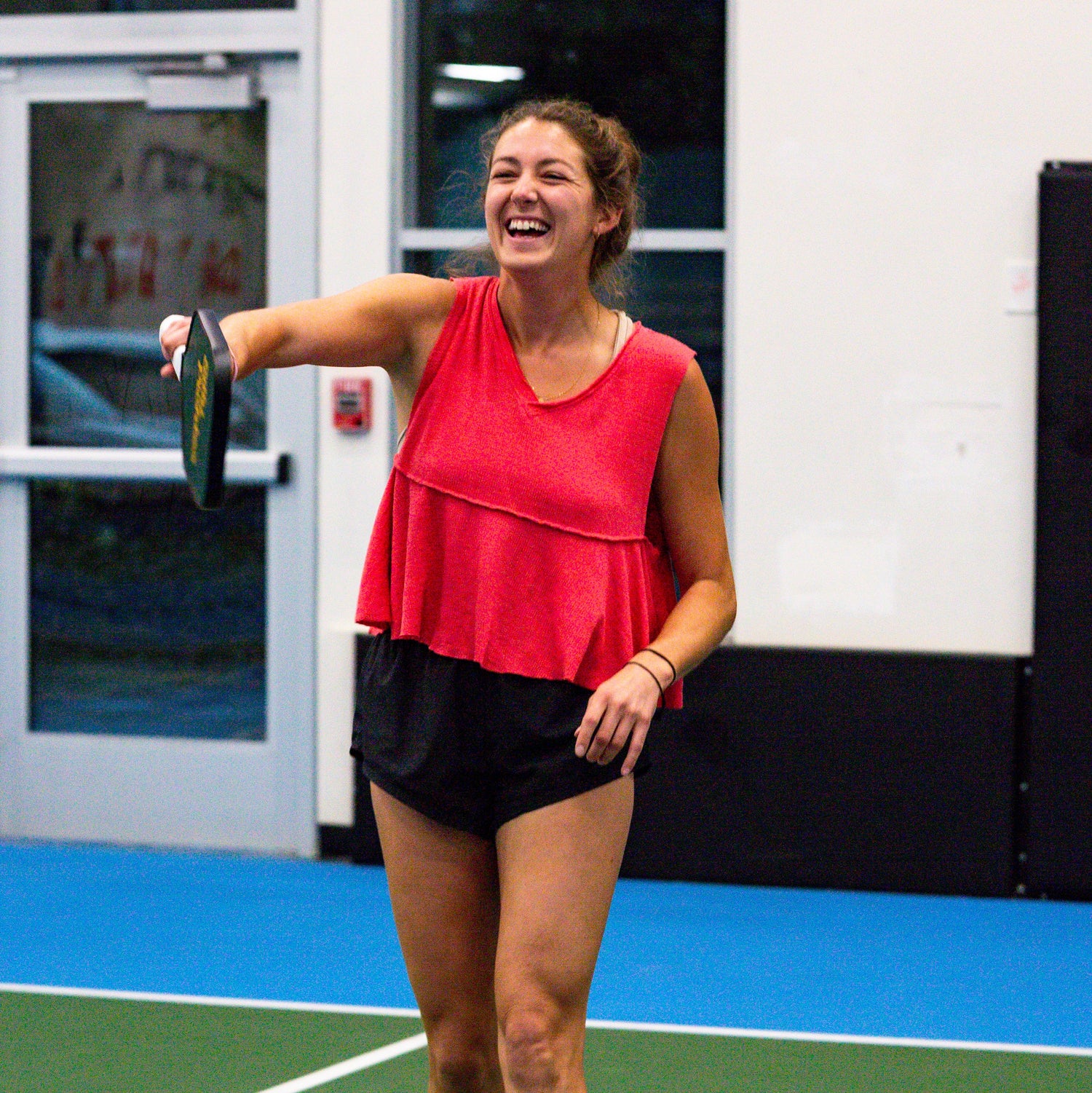 Woman in red top and black shorts laughing playing a fun game of pickleball