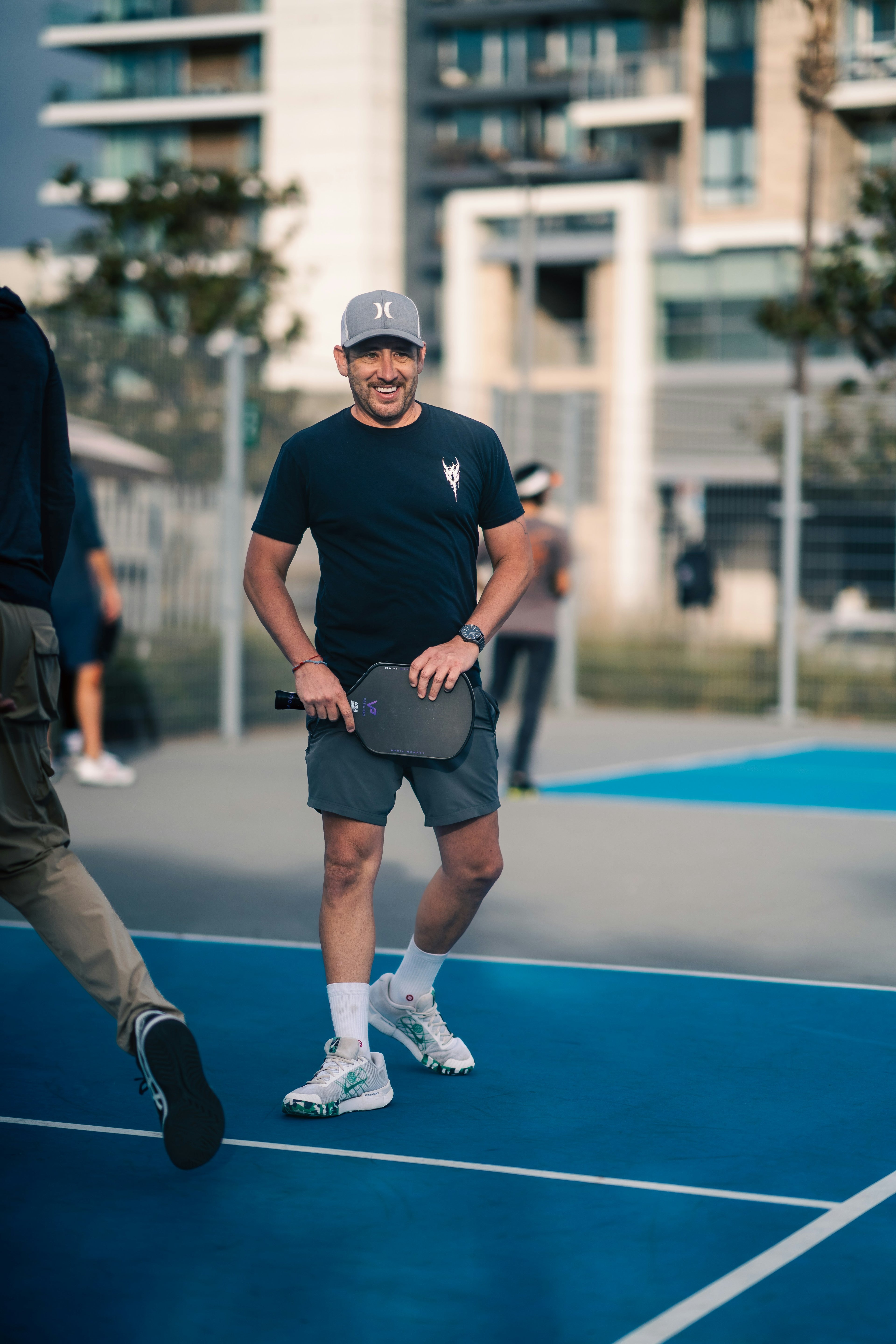 Man smiling, looking happy holding a pickleball paddle on a court with buildings in the background
